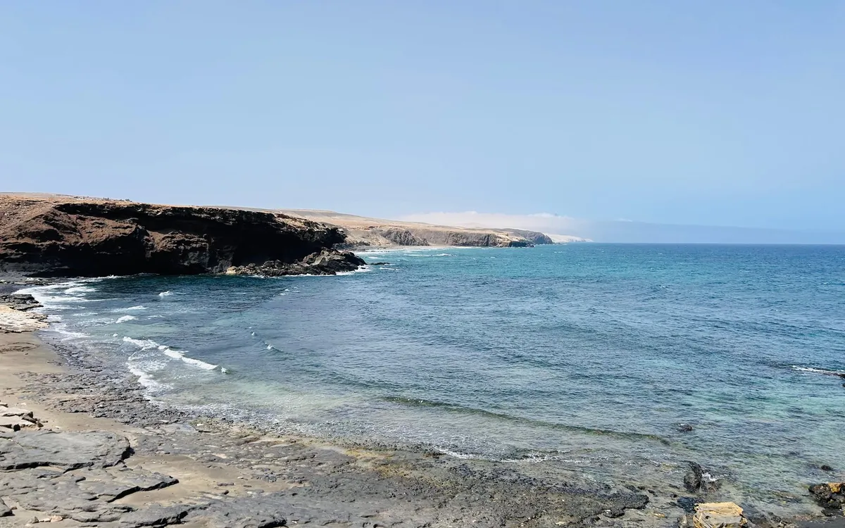 Eine kleine Bucht mit grauem Sand und flachem dunklem Fels neben klarem blauem Wasser