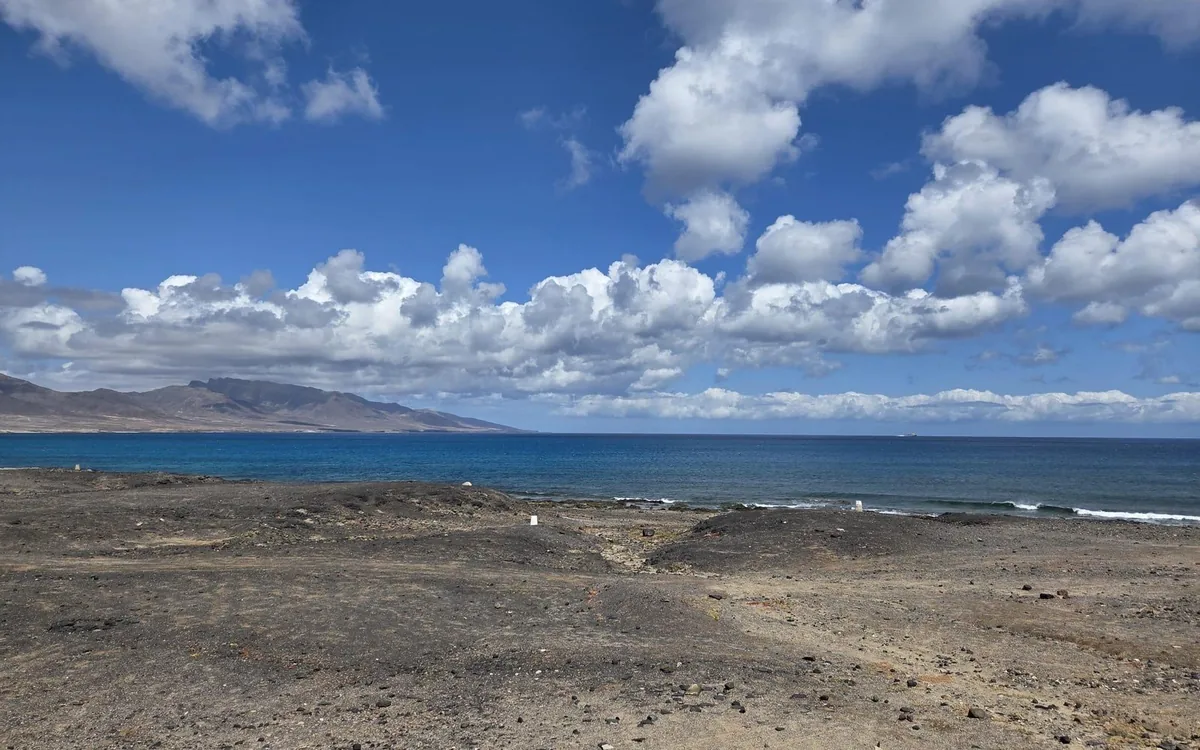 Steiniger Boden, der unter weißen Wolken zum blauen Meer abfällt, dahinter Berge