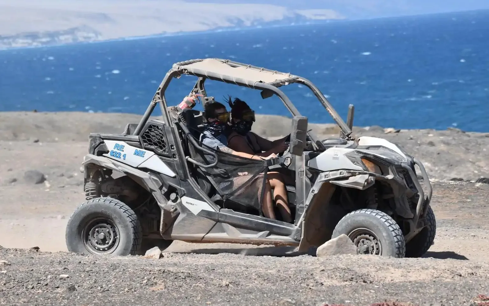 Zwei Fahrer mit Schutzbrillen und Gesichtstüchern steuern einen Buggy über steinigen Boden hoch über dem blauen Ozean